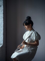 Teen girl playing the flute in a stylish white dress by the window. © Yuanxing