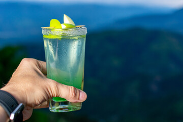 First person point of view (POV) of a hand holding a green cocktail, toasting to a vacation with a beautiful mountain view in the background.