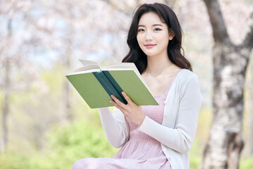 Obraz premium Young Asian female reading book outdoors in spring park, wearing pink dress and white cardigan