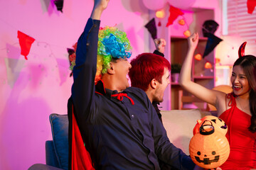 Asian friends in creative Halloween costumes gather on a sofa under colorful RGB lights, laughing and enjoying a festive night party filled with spooky decorations, joy, and playful holiday spirit.