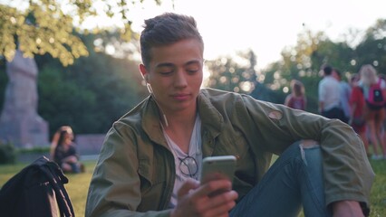 Slow motion portrait young man using smartphone with headphone sitting in park smile feel happy sunset sunlight student mobile holding handsome message boy hand Caucasian