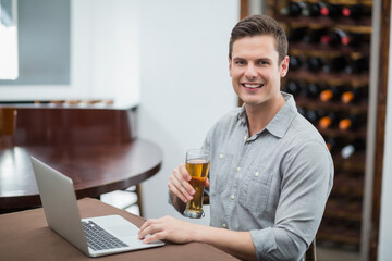 Man in late twenties typing on silver laptop holding beer glass at table in wine cellar