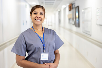 Diverse woman wearing scrubs and ID badge, standing in hospital corridor with whiteboard, smiling