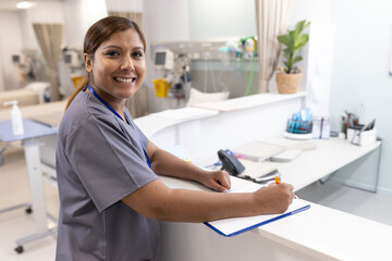 Mature adult nurse writing on clipboard with orange pen at hospital ward desk, with infusion pumps