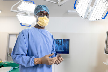 African American male surgeon donning gloves and mask in operating room with surgical lights