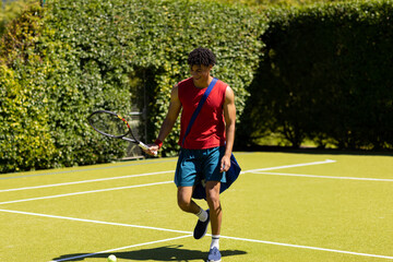 Obraz premium Young man walking on tennis court, carrying racket and blue sports bag with tennis ball