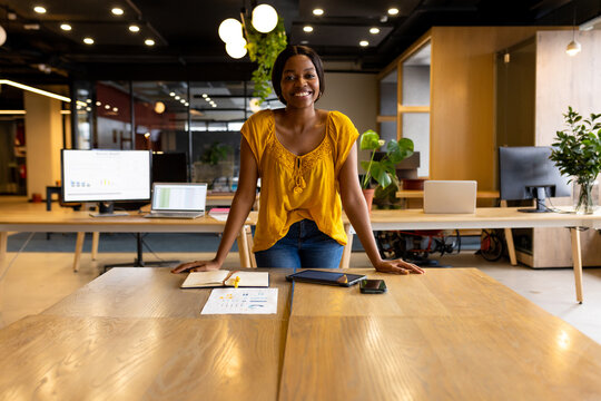 Young African American woman standing behind wooden table in modern office space, showing charts - Powered by Adobe