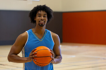 African American man standing on gym basketball court, holding basketball and smiling at camera