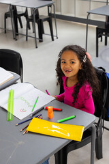 School child wearing pink shirt writing in notebook at school desk with colored pencils, copy space