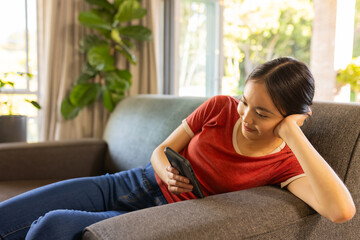 Fototapeta premium Asian woman reclining on grey sofa in living room with potted plant, holding smartphone, copy space