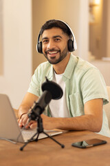 Young adult Indian man recording in home studio, typing on laptop with headphones and microphone