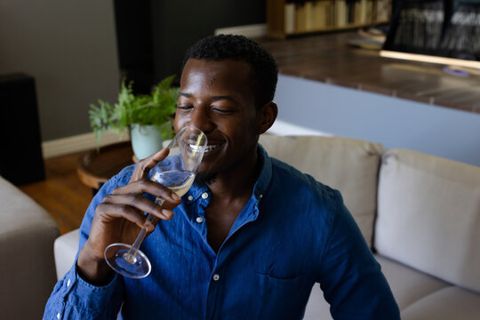 African American man drinking white wine from glass on beige couch in modern living room - Powered by Adobe