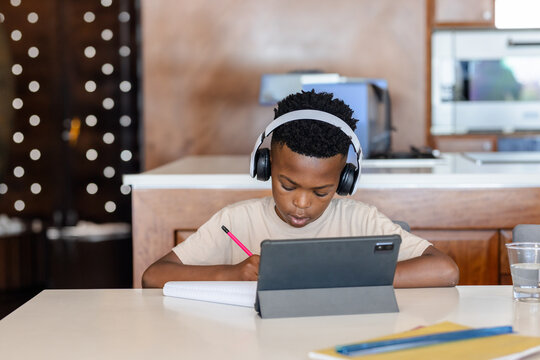 African American boy writing in notebook at kitchen table, using tablet and wearing headphones