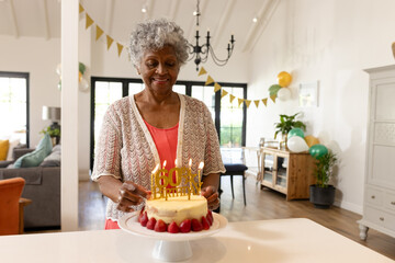 Senior woman adjusting birthday candles on cake in open-plan living room, with balloons and banner
