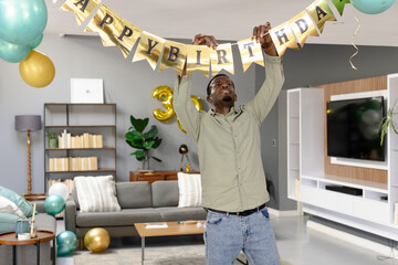 Mid-adult African American man hanging birthday banner in modern living room, with gold balloons