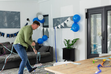 middle-aged man sweeping floor with broom in living room, clearing paper streamers and balloons