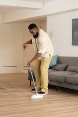 African American man sweeping colorful confetti off light wood floor in living room, with broom