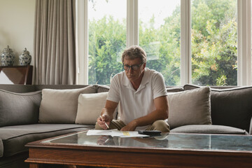 Senior man wearing polo shirt sitting in living room using highlighter on papers near smartphone