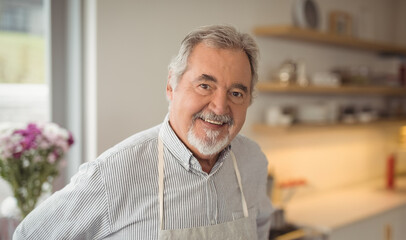 Senior man wearing striped shirt and apron smiling while standing in home kitchen with flowers