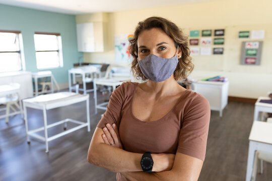 Female teacher standing with arms crossed in classroom wearing face mask beside world map poster