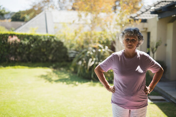Senior woman standing with hands on hips in suburban backyard by hedge and deck, copy space