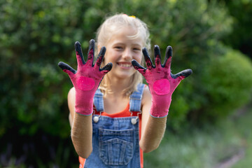 Smiling girl in garden showing dirty hands in pink gloves, enjoying outdoor fun, copy space