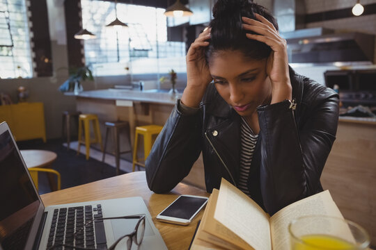 Young woman in cafe reading book, looking stressed, laptop and smartphone nearby