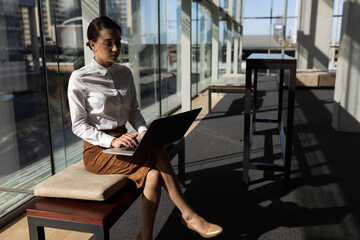Laptop resting on wooden bench near glass panels in modern office lounge, city skyline visible