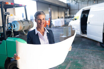 Middle-aged woman wearing blazer at warehouse holding blueprint near green forklift and cargo van