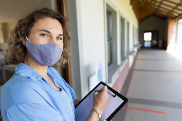 Teacher wearing mask holding clipboard, preparing for class in school hallway, copy space