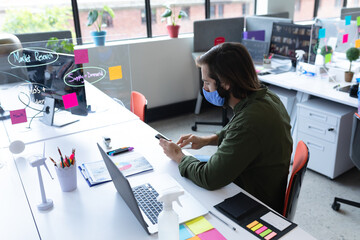 Man wearing mask using smartphone at office desk with laptop and notes