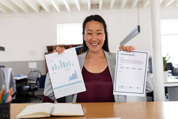 Asian woman in office holding marketing reports, smiling confidently at camera