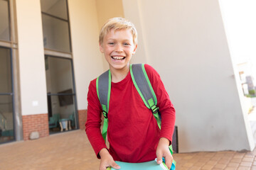 Smiling boy with backpack holding book outside school building, feeling excited