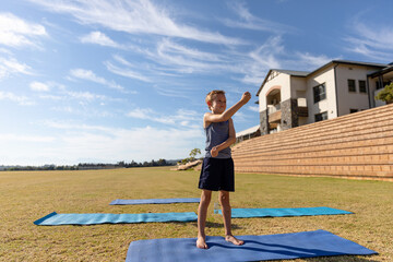 Boy stretching arms on three blue exercise mats on grassy field with wooden bleachers, copy space