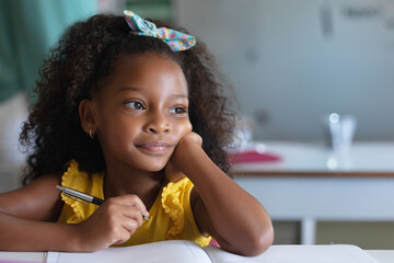 In school, young girl with pen daydreaming at desk, wearing yellow dress