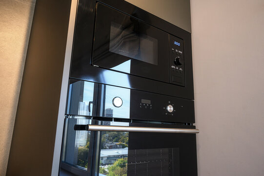 A modern, built-in black oven and microwave are stacked in a kitchen. Reflections show a cityscape outside a nearby window