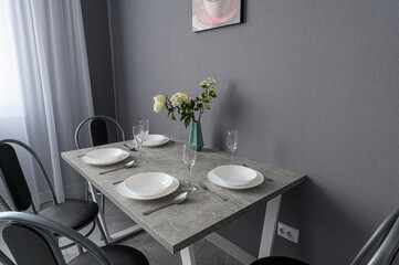 Set table with plates, glasses, and flowers on a gray table against a gray wall with black chairs