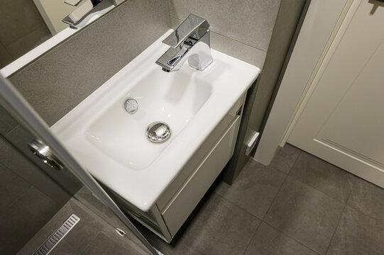A top-down shot showcases a modern bathroom sink with a chrome faucet, set against gray tiles and a white door. The vanity has a sleek, minimalist design - Powered by Adobe