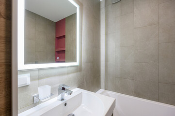 A modern bathroom featuring a sink with a chrome faucet, a light-up mirror, and tiled walls in neutral tones. Minimalist design