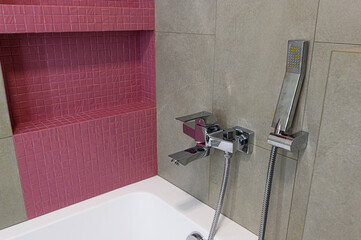 A close-up of a bathroom features a white tub with a chrome faucet and showerhead mounted on a tiled wall. A niche covered in pink mosaic tiles adds a touch of color