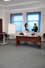 Fototapeta premium Two women in business suits discuss documents in an office near a desk and a flip chart with graphs.