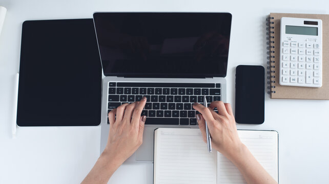 Overhead shot of business woman hand typing on laptop computer with digital tablet, smartphone and paper notebook, business document on white office desktop, business background