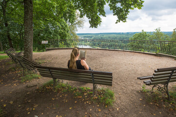 A woman with long blond hair sits on a bench and reads a book. Behind her, there is a panoramic view of the forest and the river.
