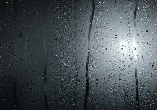 Raindrops and streaks of water on a dark, wet glass surface, with a bright light source in the background