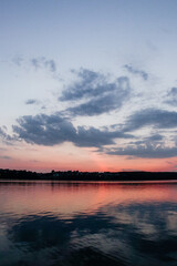 A calm lake reflects the vibrant pink and blue colors of a beautiful sunset, with the dark silhouettes of trees and buildings on the horizon.