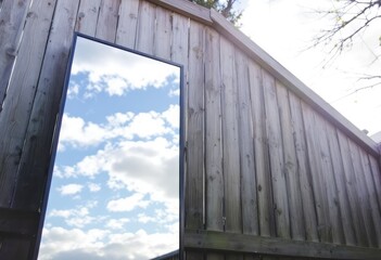 A rectangular mirror reflecting a bright, partly cloudy sky, leans against a weathered wooden fence under sunlight