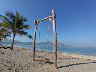 a wooden swing on the beach with a background of hills, blue sky and blue sea