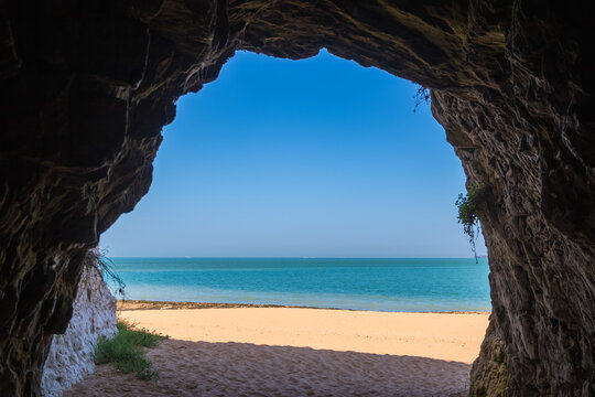 A scenic view from inside a seaside cave at Kingsgate Bay in Broadstairs, England, looking out onto a sunlit sandy beach and the calm turquoise sea under a clear blue sky