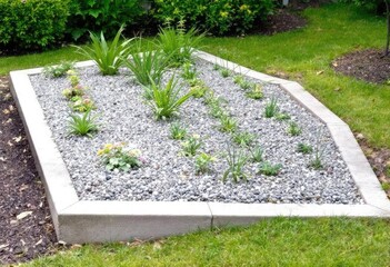 A raised, irregularly-shaped garden bed filled with gravel and various small plants, bordered by light-grey concrete edging, sits in a grassy lawn