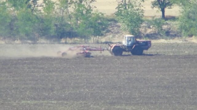 Agricultural tractor working on arid farmland in summer heat. Dust rises from soil as machinery moves across dry field. Heatwave creates shimmering mirage effect, illustrating impact of global warming